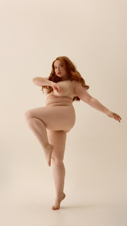 Plus-size woman in nude lingerie performing expressive dance pose on neutral background. Concept of emotional movement, body empowerment, inclusive fashion, confident self-expression.の写真素材