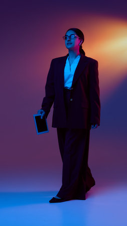 Full-body view of businesswoman walking with tablet under futuristic light. Concept of corporate branding, leadership in tech, and mobile business operations with confident ambition.の写真素材