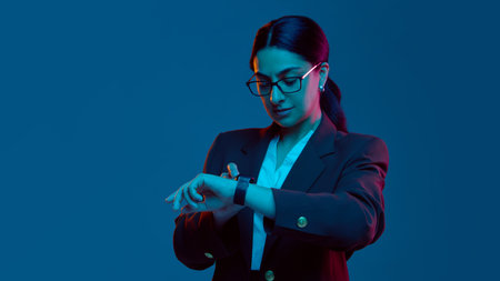 Businesswoman checking smartwatch in blue neon studio. Concept of digital lifestyle, time tracking apps, productivity tech, and corporate self-management with focused routine.の写真素材