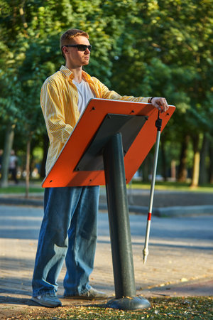 Blind man using white cane standing near accessible tactile information board in park. Concept of assistive tech, urban accessibility, social inclusion, awareness tools.の写真素材