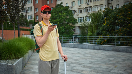 Blind man in red cap standing in city using smartphone for voice input. Concept of smart assistance,...