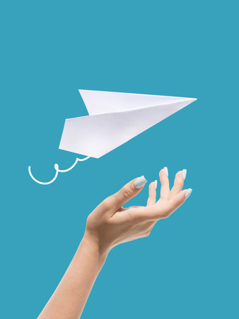 Bright collage. Female hand with manicured nails launching white paper plane on blue background with...