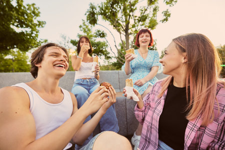 Group of friends laughing and eating street food outdoors on sunny day. Concept of street food promotion, youth lifestyle, social media content, friendship, and positive emotions.の写真素材