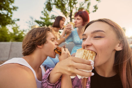 Close-up of young people enjoying burgers and wraps together in park. Concept of social dining, youth lifestyle, street food culture, friendship, and joyful urban moments.の写真素材