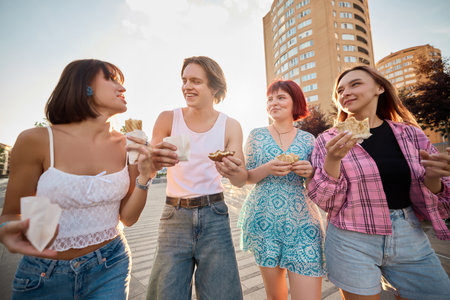 Young people walking in urban area laughing and eating fast food together. Concept of street food, lifestyle content, social media sharing, and foodtech apps.の写真素材