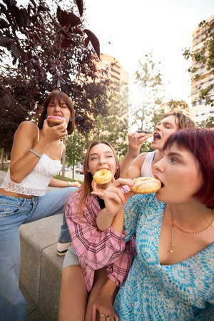 Playful young friends eating donuts and laughing together in urban outdoor setting. Concept of carefree youth, delicious street food, summer fun, and strong friendship connections.の写真素材