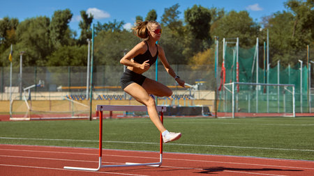 Female athlete clearing hurdle in action on stadium track. Concept of athletic competition, female empowerment, ambition, and overcoming barriers in sports and life.の写真素材