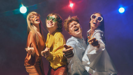 Group of friends in retro disco outfits performing playful poses under colorful lights. Concept of vintage fashion, pop culture nostalgia, joyful lifestyle, and retro-inspired branding.の写真素材
