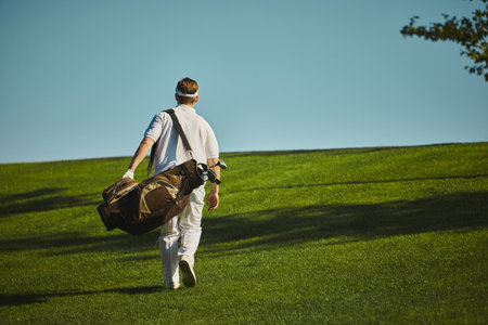 Golfer walking uphill with bag on green field under clear sky. Concept of active lifestyle, endurance, outdoor sports, leisure travel and healthy living.の写真素材