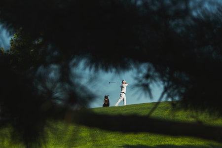 Golfer swing captured through trees in dramatic view. Concept of outdoor sport, perspective, focus, lifestyle, performance, and concentration in healthy leisure.の写真素材