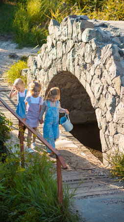 Group of little girls crossing small stone bridge holding hands and watering can. Concept of...