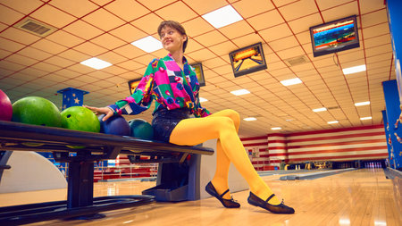 Woman sitting on bowling rack with colorful balls and playful smile. Concept of joyful sports...