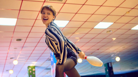 Woman in striped shirt posing playfully with bowling pin at alley. Concept of joyful lifestyle,...