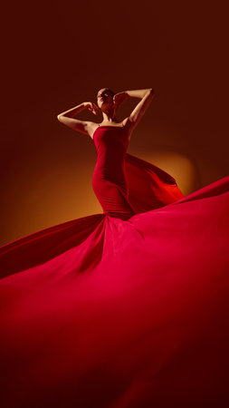 Female model in red dress posing under warm studio light with flowing fabric. Concept of fashion style, elegance, color harmony for editorial, commercial, and design projects.の写真素材