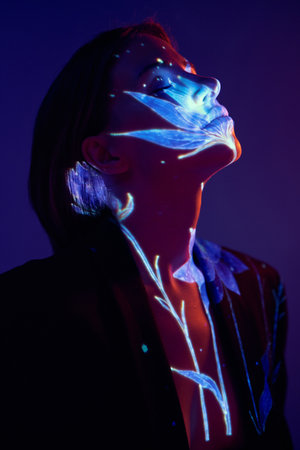 Woman with face and chest illuminated by blue floral projection in dark gradient light. Concept of emotion, identity, fusion of art and technology for editorial, design, and campaign visuals.の写真素材