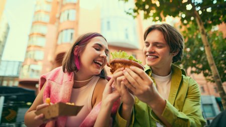 Happy young couple sharing burger and laughing outdoors together. Concept of food joy, youth lifestyle, emotional storytelling for urban marketing, social media, and food delivery visuals.の写真素材