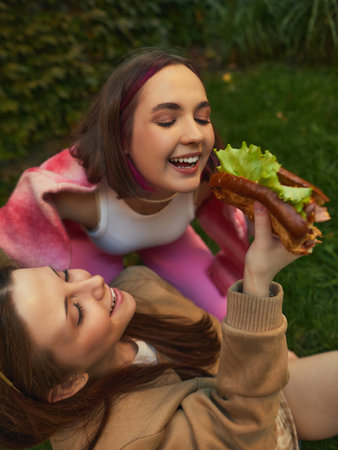Two young women laughing and enjoying burger together on grass. Concept of friendship, street-food lifestyle, emotional storytelling for youth marketing, social content, and outdoor visuals.の写真素材
