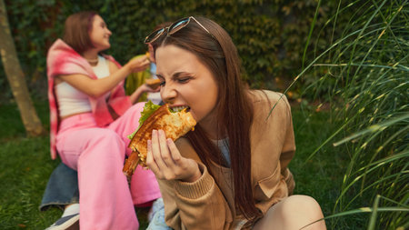 Young woman biting burger with passion while friends laugh nearby. Concept of authentic emotion, youth lifestyle, food realism for street-food branding, social content, and lifestyle marketing.の写真素材