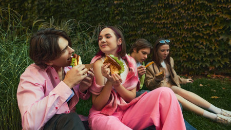 Group of young people eating burgers and fries sitting on grass together. Concept of youth friendship, outdoor lifestyle, emotional storytelling for street-food advertising and festival visuals.の写真素材