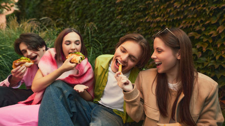 Group of friends laughing and eating burgers and fries together outdoors. Concept of youth culture, friendship, social media storytelling, and food visuals.の写真素材