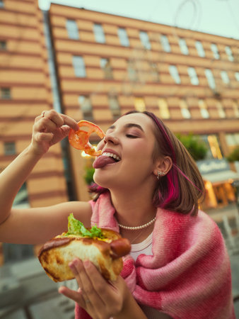 Playful young woman eating tomato slice and holding hot dog on city street. Concept of youth culture, joy, emotional expression, lifestyle advertising, and creative content.の写真素材
