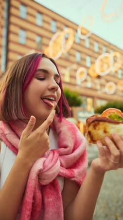 Smiling young woman tasting sauce from finger while holding hot dog outdoors. Concept of playful lifestyle, authenticity, joy for food photography, youth advertising, and urban culture campaigns.の写真素材