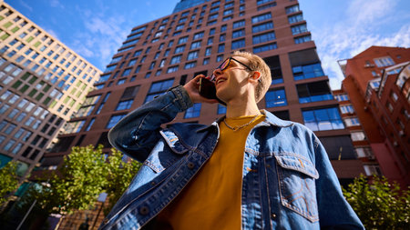 Young man in denim jacket smiling talking on smartphone among modern buildings. Concept of technology communication, lifestyle branding, marketing culture, urban professionalism.の写真素材