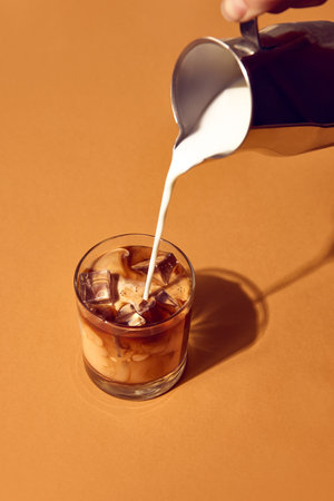 Milk pouring into iced coffee with swirling texture and glass on beige table background. Concept of barista craft, beverage advertising, product composition, and culinary lifestyle.の写真素材