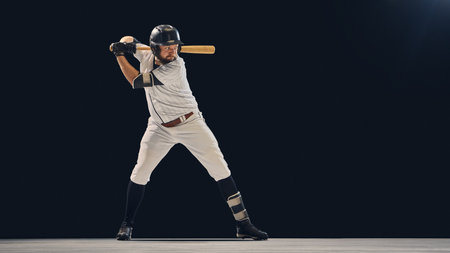 Baseball player in full uniform holding bat in batting stance on dark background. Concept of...