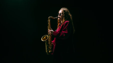 Female musician standing with saxophone in dark atmosphere with cinematic light. Concept of creative advertising, music streaming visuals, and psychology of confidence and focus.の写真素材