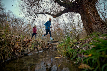 Male and female runners sprinting across forest ridge covered in fallen autumn leaves. Concept of sports performance, fall outdoor content, athletic discipline and active lifestyle promotion.の写真素材