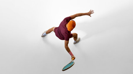 Male tennis player in orange cap reaching wide forehand from overhead angle on white floor. Concept of creative advertising, visual direction, storytelling movement, and athletic inspiration.の写真素材
