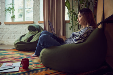 Woman working on laptop in cozy home interior with warm daylight. Concept of business lifestyle, remote work visuals, productivity routines, informal workspace ads, and creative working moods.の写真素材