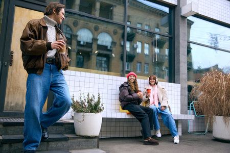 Man standing with coffee while friends sit and chat outdoors. Concept of winter tourism, social content, event posters, warmth and mobile delivery services.の写真素材