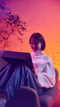 Woman smiling while using tablet under warm neon gradient light. Concept of digital engagement, online communication, creative workflow, lifestyle content for courses, blogs, apps and social platformsの写真素材