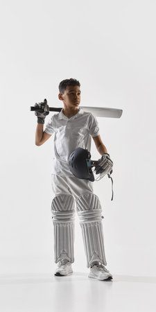 Young cricket player standing with bat on shoulder holding helmet in studio. Concept of youth sport training, athletic lifestyle, equipment advertising and cricket academy promotion.の写真素材