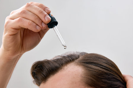 Close up of mans head with hand applying serum to hair with dropper on light gray background in...