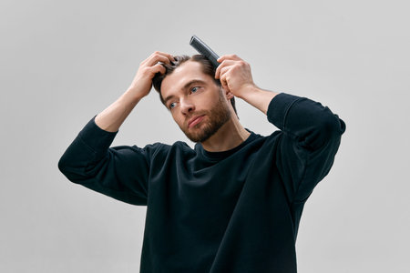 Adult short bearded man in black sweatshirt combing hair with black comb while holding hair with...