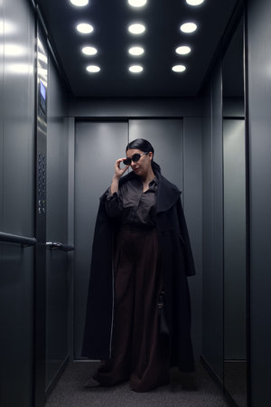 Elegant woman adjusting sunglasses while standing inside modern elevator wearing dark coat. Concept...