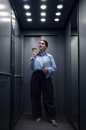 Young professional woman holding coffee cup and laptop inside modern elevator. Concept of busy...