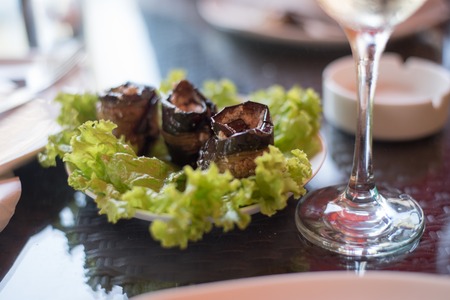 A dish of vegetable snack rolls of stuffed eggplant served with green salad leaves and a glass of wine on a table in a restaurantの写真素材