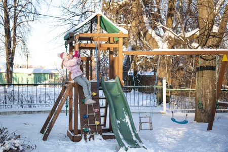A cheerful child girl plays on the Playground on a snowy winter bright sunny frosty dayの写真素材