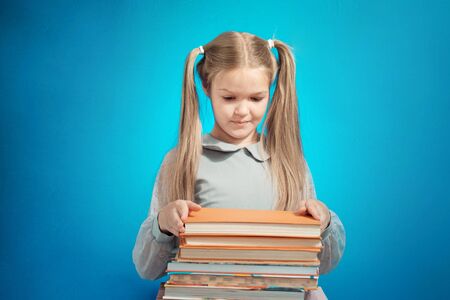 Cute Caucasian schoolgirl in surprise looks at a big on a stack of books on a blue background....