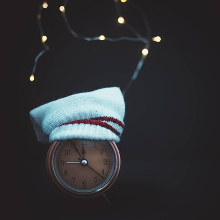 Close up of retro alarm clock in a knitted white hat on dark background with bokeh Christmas lightの写真素材
