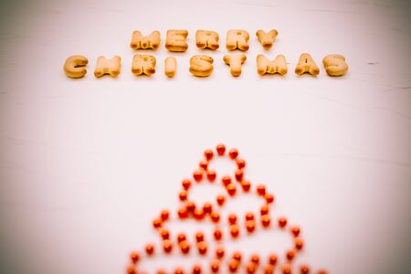 The words merry christmas of cookie letters. Flat lay composition for greeting card om white background. Blurred christmas tree made red beads, toned photo, horizontal formatの写真素材