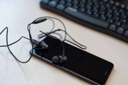 Flat lay composition with headphone and black smartphone in the foreground, against a blurred background with a computer keyboard.On begie desk backgroundの写真素材