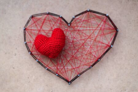 Small knitted heart lies on big red heart made of threads, stranded between the nails. The concept of great nascent or hidden love.の写真素材