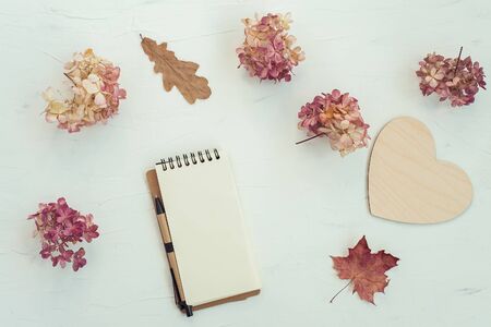 Workspace with notebook on white background. Autumn composition with wooden heart, dry flowers and leaves. Flat lay, top view, copy space.の写真素材