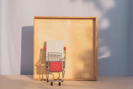 Grocery mini trolley cart with white card casting a shadow on wooden frame. front view, copy space, template.の写真素材