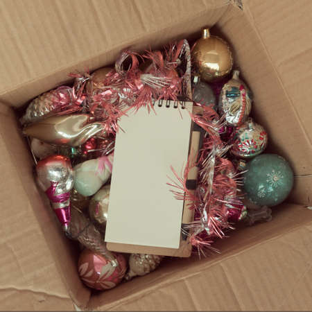 An open cardboard box with old glass Christmas decorations. On top of a clean notepad with a ball...